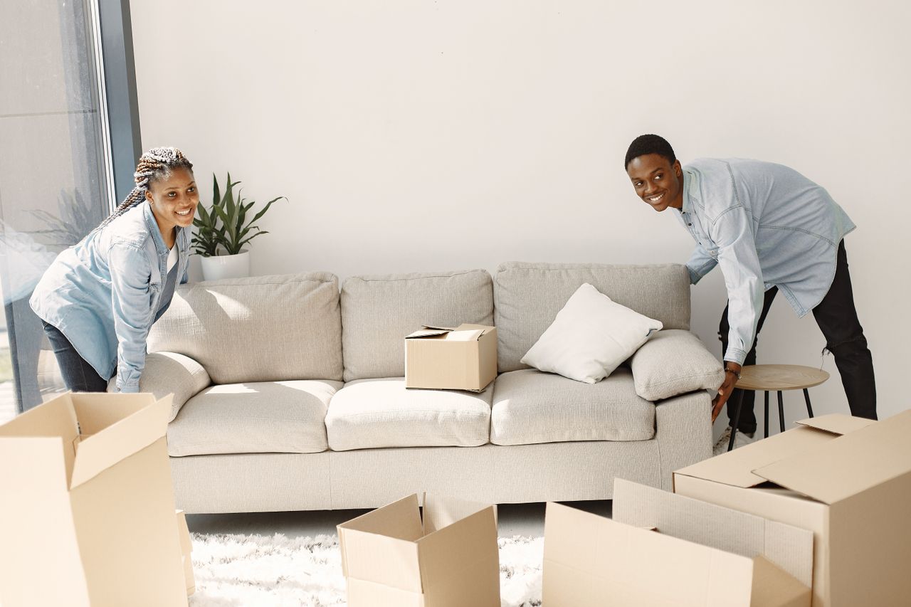 Un couple souriant déplaçant des meubles dans un salon moderne et lumineux, entouré de cartons.